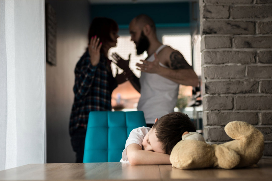 A boy sitting at a desk, while a woman and a man are arguing in the background.