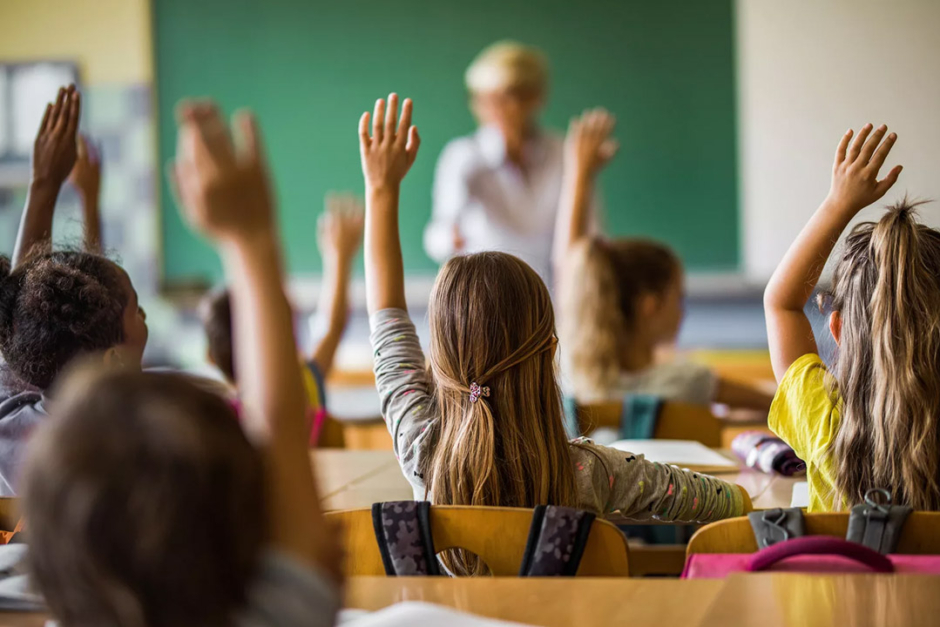 Children in classroom with raised hands, in front of a blurred teacher in the background.