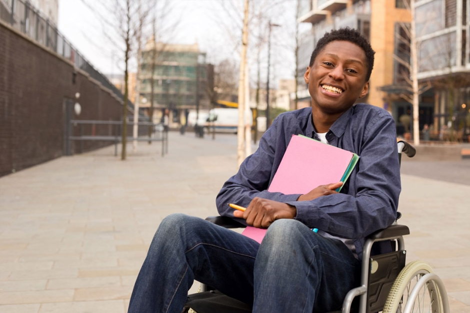 A person in a wheelchair in the street, holding a pen and a stack of papers.