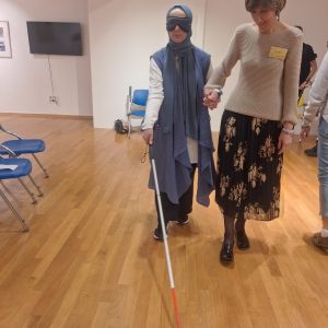 A woman with a headscarf and an eyeband walking with a white cane, alongside a sighted person.