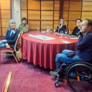 A group of people, sitting around a table talking, one of them in a wheelchair.
